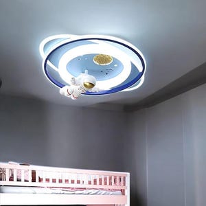 May include: A circular ceiling light fixture in a planet design, featuring blue and white concentric rings. A small astronaut figure hangs below. The light includes a gold-coloured planet and star details.