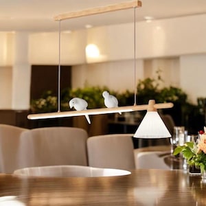 May include: A wooden pendant light fitting with a white shade and three white bird figurines perched on a wooden branch. The light fitting is hanging over a wooden table.