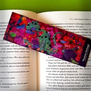 Könnte beinhalten: Ein pinkfarbenes und grünes Lesezeichen mit einem Comic-Dinosaurier und einer Katze. Das Lesezeichen trägt den Text "Thomas Hufnagl" in der rechten unteren Ecke.