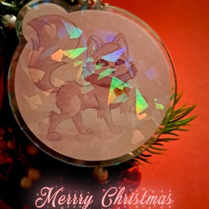 Könnte beinhalten: Runder, klarer Weihnachtsschmuck mit einer Cartoon-Fuchs-Illustration. Der Fuchs ist hellrosa mit einer roten Fliege. Der Schmuck hat holografische Akzente und die Worte "Merry Christmas" in Rosa.