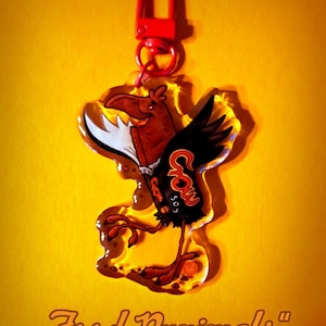 Könnte beinhalten: Ein klarer Acryl-Schlüsselanhänger mit rotem Verschluss. Der Schlüsselanhänger zeigt ein Cartoon-Vogel-Design mit einem Schokoriegel-Körper und schwarzen Flügeln. Der Text "Food Punimals" befindet sich unten.