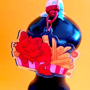 May include: A clear acrylic keychain featuring a red cartoon character and french fries in a striped container. The keychain has a red clasp and a small logo in the corner. The background is a solid yellow.