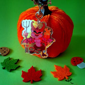 Könnte beinhalten: Ein Halloween-Schlüsselanhänger mit einem Cartoon-Teufel, der an einem orangefarbenen Kürbis hängt. Der Kürbis ist von Filzblatt- und Pilzdekorationen umgeben. Der Satz "HAPPY HALLOWEEN" ist in Rot geschrieben.