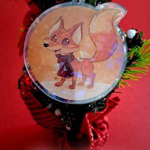 Könnte beinhalten: Runder Weihnachtsschmuck mit einem Cartoon-Fuchs, der einen Schal trägt, vor einem schneebedeckten Hintergrund. Der Schmuck hat einen holografischen Effekt und ist an einer goldfarbenen Kette befestigt. Die Worte "Merry Christmas" stehen unten.