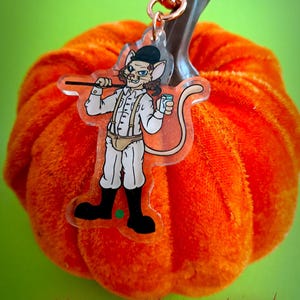 May include: An orange plush pumpkin with a clear acrylic charm featuring a cartoon cat in a white outfit, black boots, and a black hat. The charm is attached to a gold-colored clasp. The words "HAPPY HALLOWEEN" are written in red at the bottom.