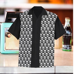 Puede incluir: Una camisa negra de manga corta con botones y un patrón de calaveras. La camisa tiene un diseño dividido con un panel negro sólido en el centro.