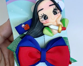 May include: A set of hair accessories featuring a cartoon character. The set includes a blue ribbon clip with a heart and gem, and a red and blue bow. The character has black hair and is holding a sword. The background is a light blue card with a rainbow.