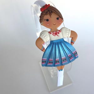May include: A blue and white headband with a handmade doll figure wearing a blue skirt, white blouse, and red bow. The doll has brown hair and a smiling face.