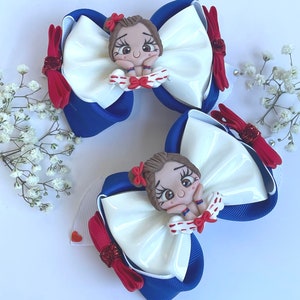 May include: Two white and blue hair bows with red accents. Each bow features a small, hand-painted doll with brown hair, brown eyes, and a red and white dress.