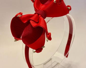 May include: A vibrant red headband featuring a large bow and decorative rose accents. The headband is made of a glossy ribbon material and is mounted on a clear plastic frame. The bow is approximately 6 inches wide.