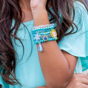 May include: A stack of blue and clear beaded bracelets with a gold charm featuring a cartoon girl's face. The bracelets are worn on a person's wrist.