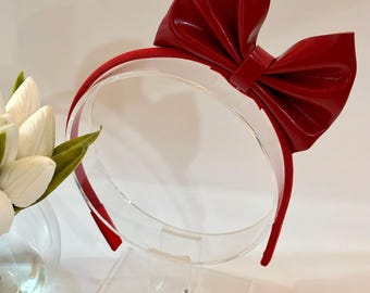 May include: A red headband with a large bow. The bow is made of a shiny, patent-like material. The headband is displayed on a clear, acrylic stand. A vase of white flowers is visible in the background.