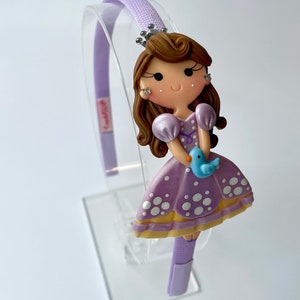 Pode incluir: Faixa de cabelo roxa com um vestido roxo e branco de bolinhas, cabelo castanho e uma coroa. A boneca está segurando um pássaro azul.