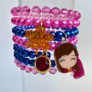 May include: Stack of beaded bracelets in shades of pink and blue. The bracelets have charms, including a gold glitter star, a red gem, and a cartoon girl with a pink tassel. The bracelets are stacked on a clear glass.
