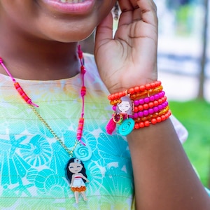 May include: A young girl wearing a colorful necklace and bracelet featuring a cartoon character. The necklace has a gold chain with a turquoise swirl charm and a pendant of a cartoon girl with brown hair wearing a pink and orange dress. The bracelet has multiple layers of orange, pink, and gold beads with a turquoise swirl charm and a pendant of a cartoon girl with brown hair wearing a pink and orange dress.