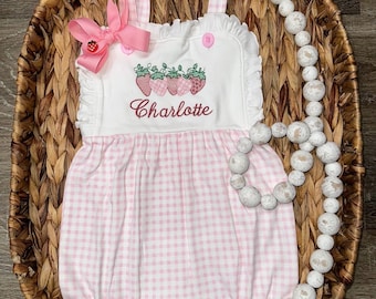 May include: A white and light pink gingham romper featuring a ruffled neckline, two pink buttons, and an embroidered row of strawberries above the name Charlotte in red script. A pink bow with a small strawberry charm is attached to the strap.