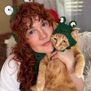 May include: A dark green crochet hat featuring two protruding frog eyes with white and black centers. The hat is designed with a textured, open-stitch pattern and includes ear flaps that tie under the chin, mimicking the appearance of a frog head.