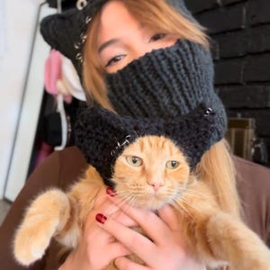 May include: A black knit cat ear hat with silver accents is worn by an orange tabby cat. The cat is being held by a person wearing a matching black knit face covering. The cat's eyes are open, and it appears to be looking at the camera.