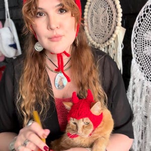 May include: A red crocheted devil horn hat is worn by a person and a cat. The person is holding a crochet project with a red yarn. The cat is orange with a red hat. The person is wearing a black top and jewelry.