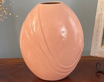May include: A pink ceramic vase with a smooth, glossy finish and a unique, curved design. The vase is sitting on a wooden surface.