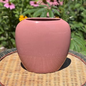 May include: A smooth, rounded, dusty pink ceramic vase with a wide, open top and a tapered base. The vase features a glossy, reflective finish that catches the light, creating a clean, simple silhouette without any additional patterns or textures.