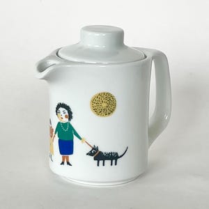 May include: White ceramic creamer with a lid, featuring a whimsical illustration of a person walking a black dog. A yellow sun-like design is also present. The creamer has a handle and a spout.
