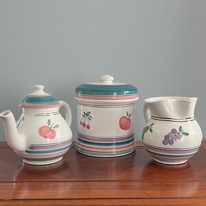 May include: A set of three ceramic kitchenware items with a fruit motif. A teapot, a canister, and a pitcher are decorated with pink apples, green cherries, and purple grapes.