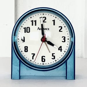 May include: A blue retro-style alarm clock with a white face and black numerals. The clock has a clear, blue plastic frame and a red second hand. The word "ADVANCE" is printed on the clock face.