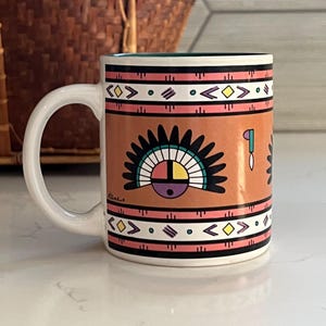 May include: A ceramic mug with a white handle and a brown body. The mug features a Native American-inspired design with geometric patterns in black, pink, yellow, and white. The design includes a sun symbol and other stylized shapes.