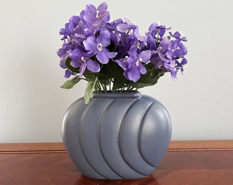 May include: A gray ceramic vase with a wavy design, holding a bouquet of purple flowers.