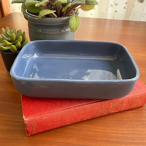 May include: A blue ceramic rectangular baking dish sitting on top of a red book. The dish is empty and has a glossy finish.