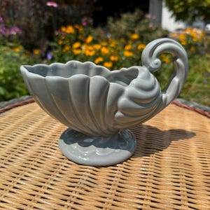 May include: A glossy light gray ceramic gravy boat or sauce server featuring a scalloped shell-like design, a wide fluted bowl, a pedestal base, and a single curved handle that loops upward and attaches to the side of the vessel.
