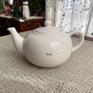 May include: A white ceramic teapot with a rounded body, a curved spout, and a loop handle. The lid has a small knob. The word "tea" is printed in black on the front. The teapot is on a textured, light-coloured surface.