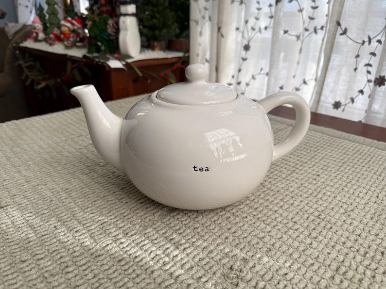 May include: A white ceramic teapot with a rounded body, a curved spout, and a loop handle. The lid has a small knob. The word "tea" is printed in black on the front. The teapot is on a textured, light-coloured surface.