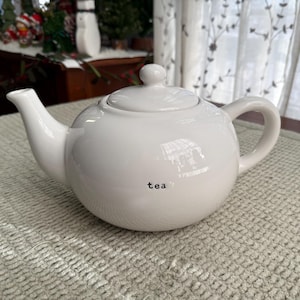 May include: A white ceramic teapot with a lid and handle. The word "tea" is printed in black on the side. The teapot is on a light green textured surface.
