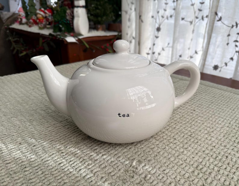 May include: A white ceramic teapot with a lid and handle. The word "tea" is printed in black on the side. The teapot is on a light green textured surface.
