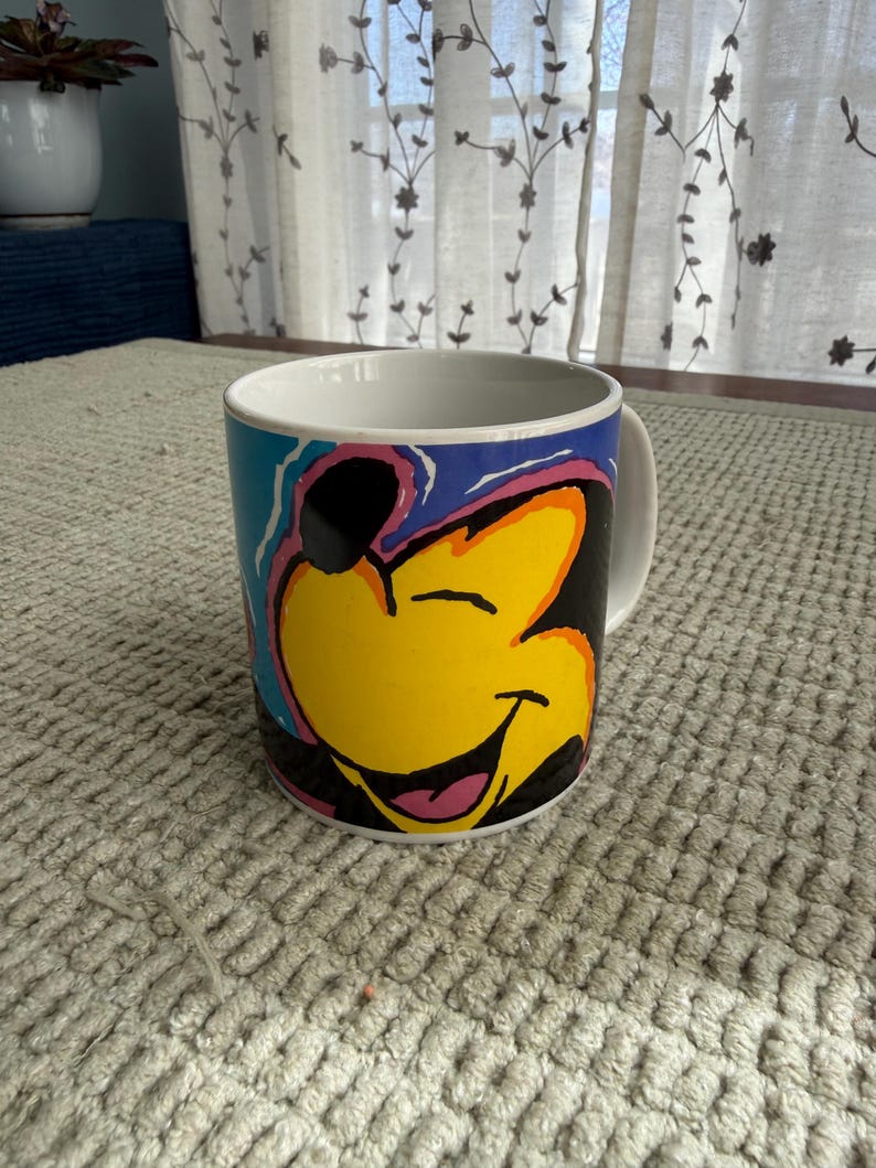 Peut inclure: Mug en c&eacute;ramique blanche avec un personnage de dessin anim&eacute; souriant. Le personnage a un visage jaune, des oreilles noires et une langue rose. Le mug a un fond bleu avec des accents color&eacute;s. Une anse blanche est visible sur le c&ocirc;t&eacute; droit.