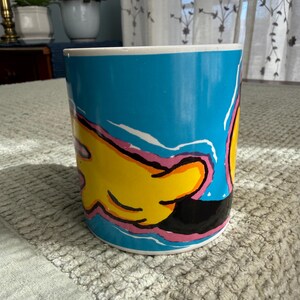 Peut inclure: Une tasse &agrave; caf&eacute; en c&eacute;ramique avec un fond bleu et un motif de main de dessin anim&eacute; en jaune, noir et rose. La tasse a un bord blanc et est de forme cylindrique. La tasse est pos&eacute;e sur une surface textur&eacute;e gris clair.