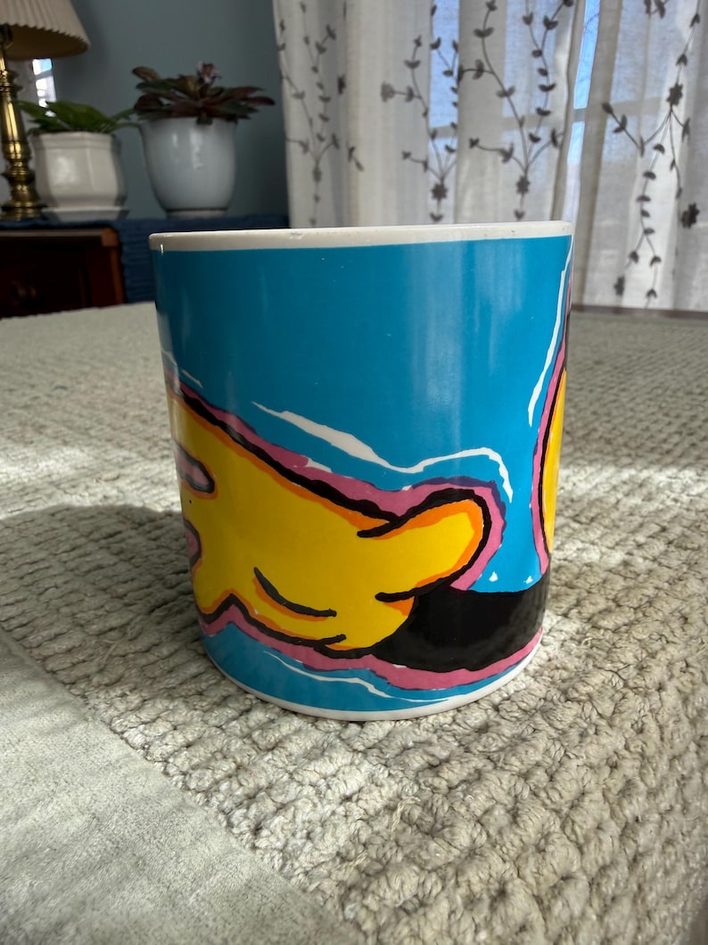 Peut inclure: Une tasse &agrave; caf&eacute; en c&eacute;ramique avec un fond bleu et un motif de main de dessin anim&eacute; en jaune, noir et rose. La tasse a un bord blanc et est de forme cylindrique. La tasse est pos&eacute;e sur une surface textur&eacute;e gris clair.