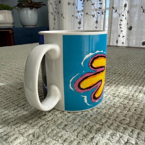 Peut inclure: Mug en c&eacute;ramique blanche avec une grande anse incurv&eacute;e. Le mug pr&eacute;sente un motif vibrant d'une fleur jaune avec des accents roses et bleus sur un fond bleu vif. Le mug est sur une surface textur&eacute;e.