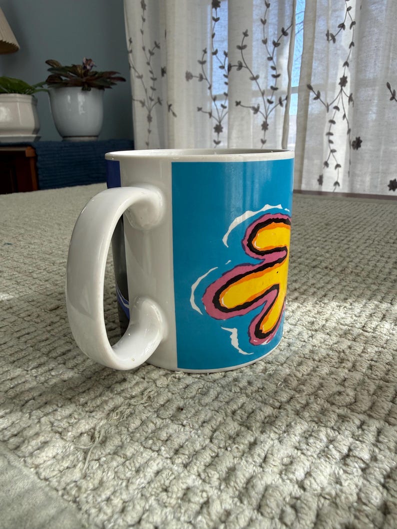 Peut inclure: Mug en c&eacute;ramique blanche avec une grande anse incurv&eacute;e. Le mug pr&eacute;sente un motif vibrant d'une fleur jaune avec des accents roses et bleus sur un fond bleu vif. Le mug est sur une surface textur&eacute;e.