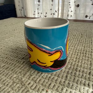 Peut inclure: Mug en c&eacute;ramique avec un fond bleu et un motif de main jaune. La main a des contours roses et noirs. L'int&eacute;rieur du mug est blanc et il repose sur une surface textur&eacute;e de couleur claire.
