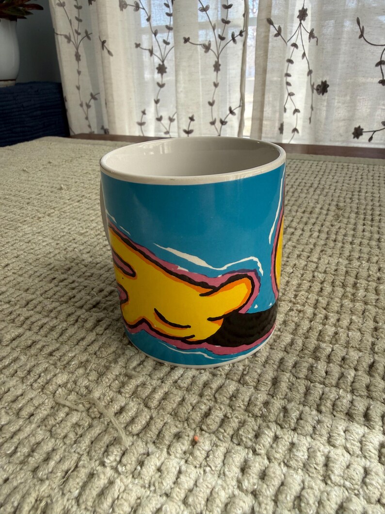 Peut inclure: Mug en c&eacute;ramique avec un fond bleu et un motif de main jaune. La main a des contours roses et noirs. L'int&eacute;rieur du mug est blanc et il repose sur une surface textur&eacute;e de couleur claire.