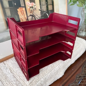May include: A stackable four-tier desk organizer in a deep burgundy red color. This office supply unit features four open-front trays with ribbed side panels and integrated rectangular handle cutouts on each side for easy access to documents.