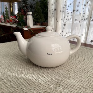 May include: A white ceramic teapot with a rounded body, a curved spout, and a loop handle. The word "tea" is printed in black on the front. The teapot is on a textured, light-coloured surface.