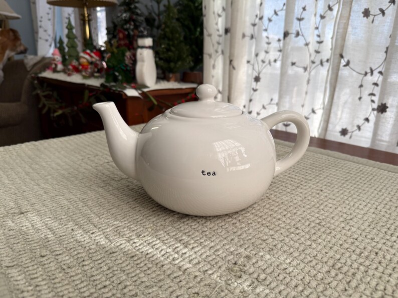 May include: A white ceramic teapot with a rounded body, a curved spout, and a loop handle. The word "tea" is printed in black on the front. The teapot is on a textured, light-coloured surface.