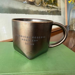 May include: A bronze-colored ceramic mug with a square base and a rounded handle. The mug has the text "STARBUCKS RESERVE ROASTERY CHICAGO" printed on the side. The mug is sitting on a green surface.