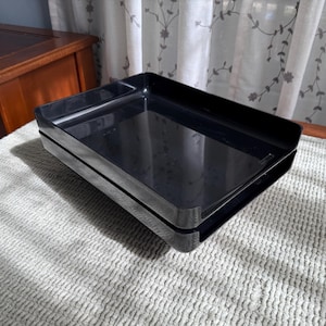 May include: Two black rectangular trays stacked on top of each other. The trays are made of a glossy material and have a simple, functional design. The trays are sitting on a textured, light-colored surface.