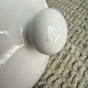 May include: Close-up of a white ceramic lid with a rounded knob. The lid has a smooth, glossy finish, and the knob is a perfect sphere. The background is a textured, light-coloured surface.