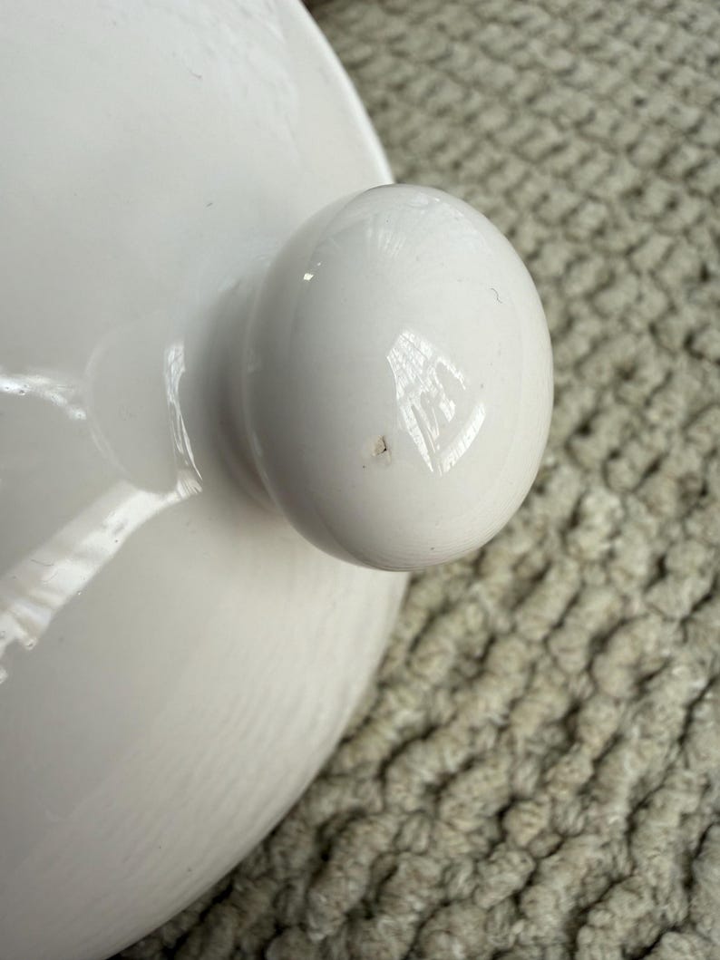 May include: Close-up of a white ceramic lid with a rounded knob. The lid has a smooth, glossy finish, and the knob is a perfect sphere. The background is a textured, light-coloured surface.